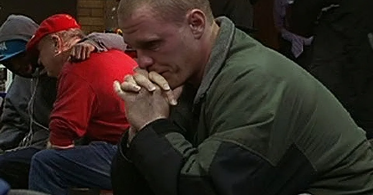 A man in a green jacket sits with his hands clasped, appearing contemplative or distressed.