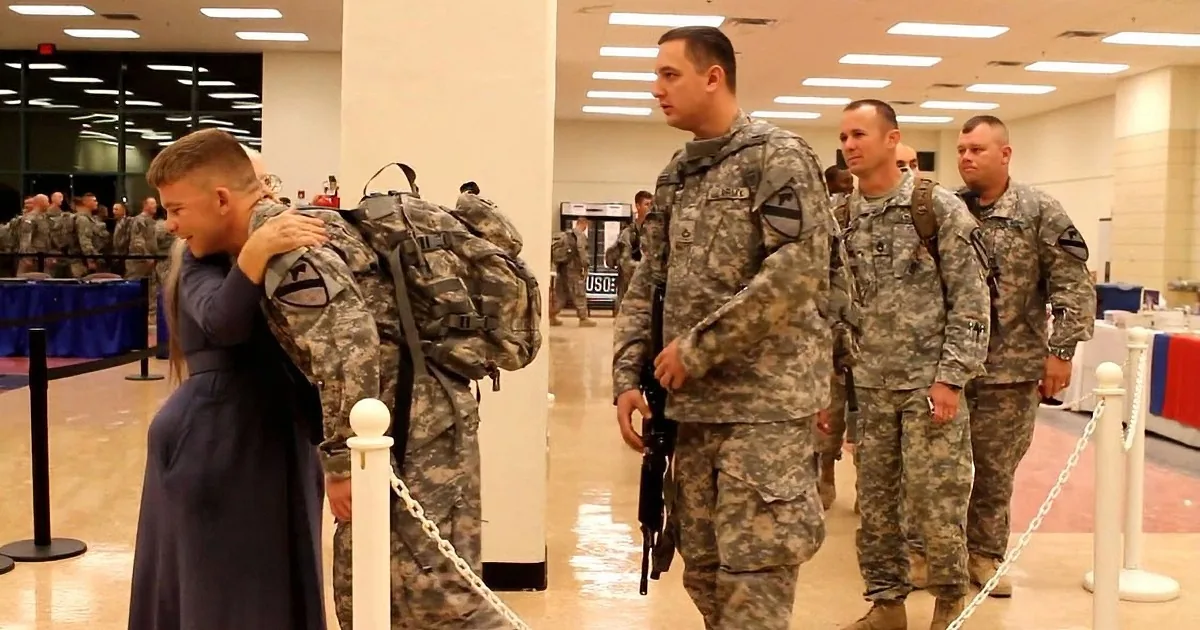 A soldier in uniform hugs a child while other soldiers walk in a line, carrying backpacks.
