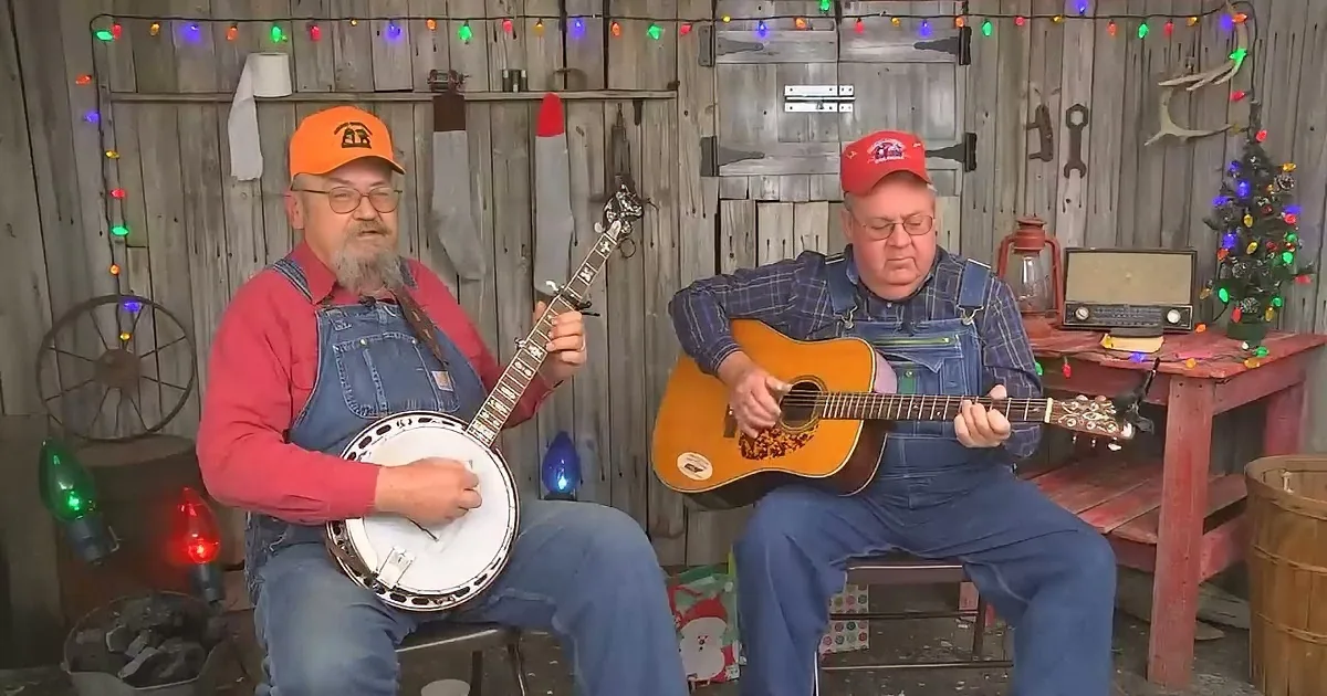 Two men play banjo and guitar in a rustic setting with Christmas lights.