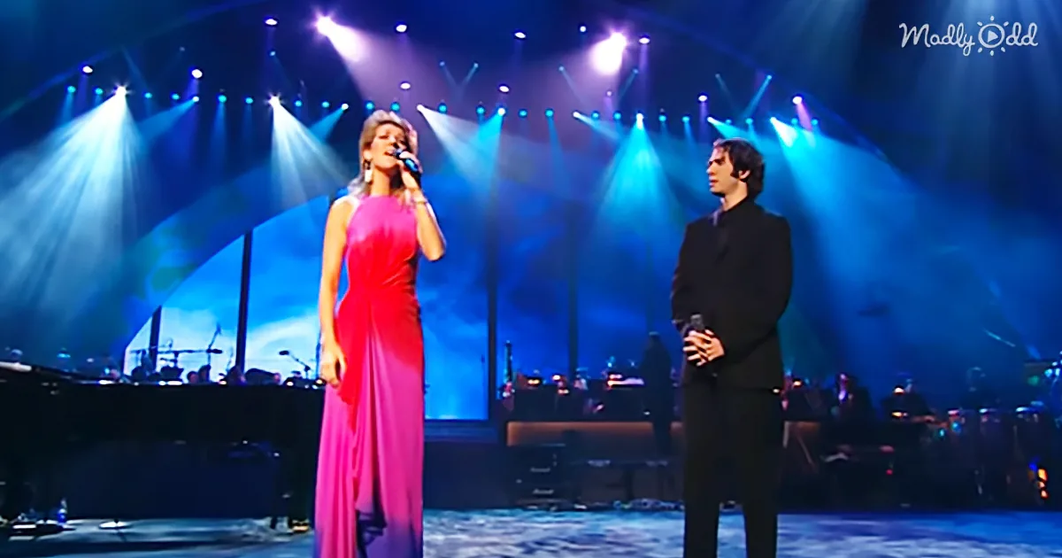 A woman in a pink dress sings on stage, accompanied by a man in a suit, under bright stage lights.