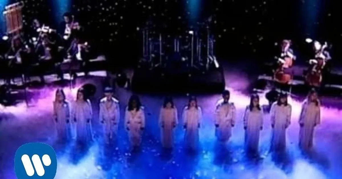 A group of performers in white outfits stands on a stage with a band and blue lighting.