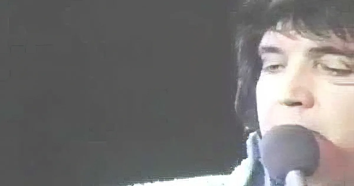 A person is singing into a microphone, wearing a dark shirt, with a blurred background.