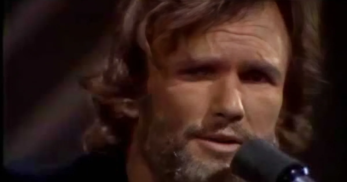 A man with long hair and a beard is speaking into a microphone, partially obscured by the microphone.