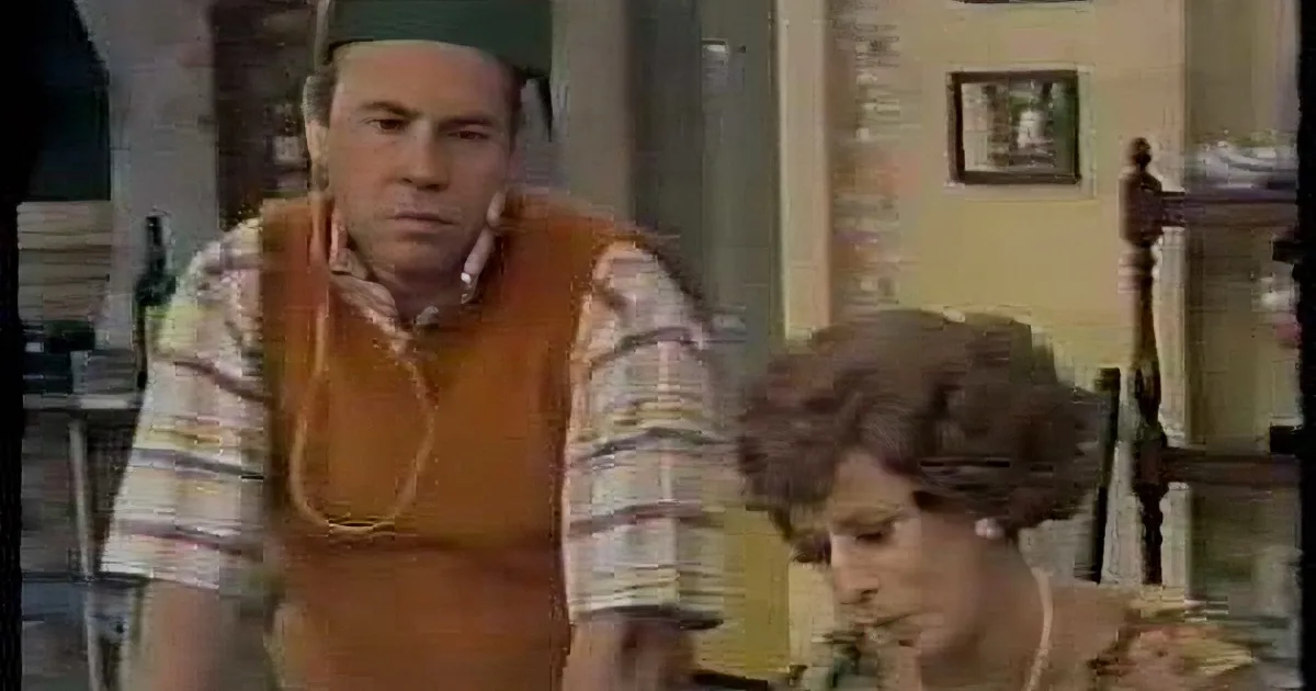 A man in a striped shirt and orange vest sits at a table, looking to his left, with a woman in a floral dress beside him.