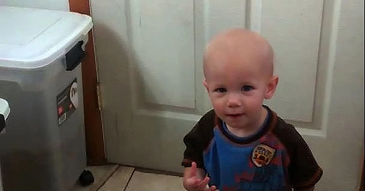 A young child with a bald head wearing a blue and brown shirt stands indoors near a white door and a gray storage bin.