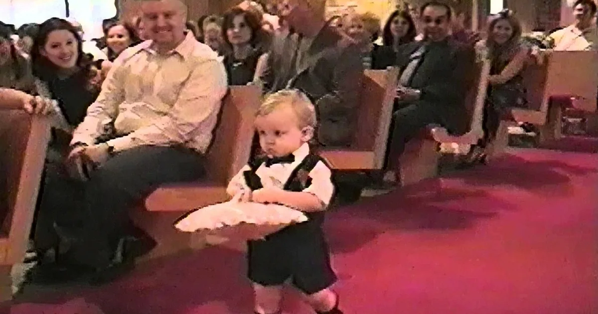 A toddler in a tuxedo sits on a chair, surrounded by adults in a church setting.