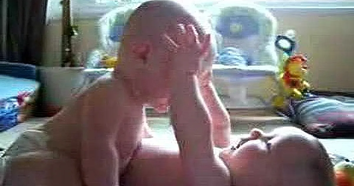 A baby is sitting on the floor, covering its face with its hands, surrounded by toys and a window in the background.