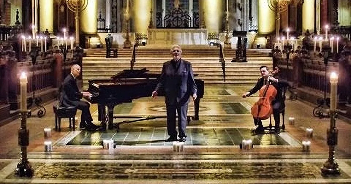 A man stands in a grand hall with a piano, surrounded by candles and a violinist in the background.