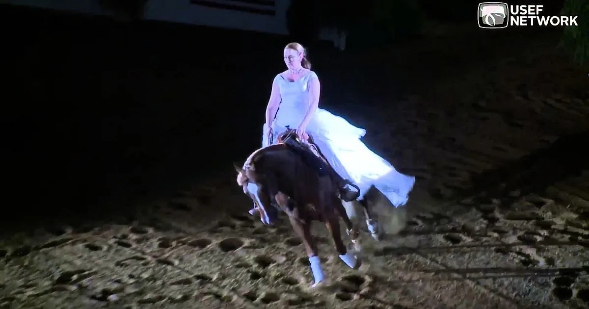 A woman in a white dress rides a horse in a dimly lit arena, her dress flowing behind her.