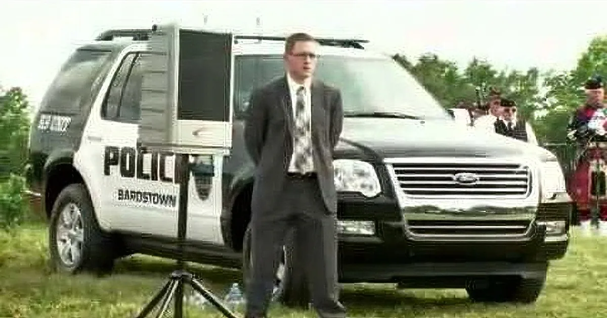 A man in a suit stands in front of a police SUV, with other vehicles and people in the background.