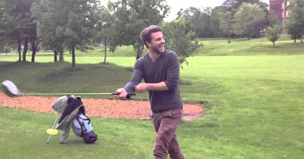A man in a park smiles while holding a golf club, with a bag and another person in the background.