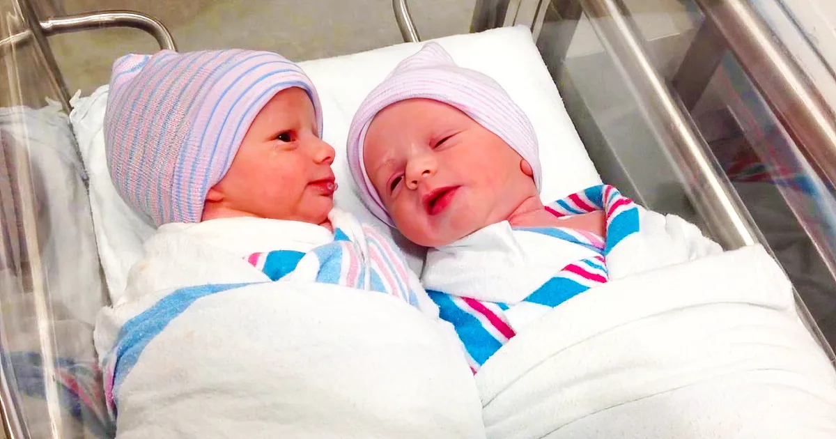 Two newborn babies wrapped in blankets and wearing striped hats rest side by side in a hospital bassinet.