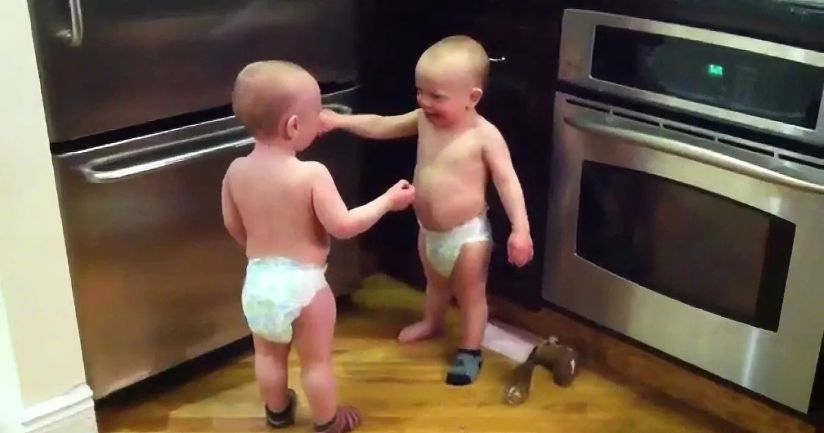 Two toddlers, one in a diaper, interact near a stainless steel oven in a kitchen.