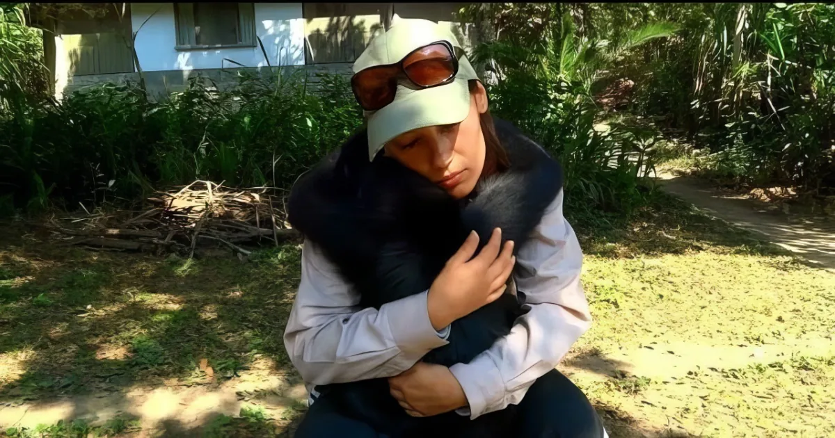 A person wearing a cap and sunglasses holds a black animal, possibly a cat, in a grassy outdoor setting.