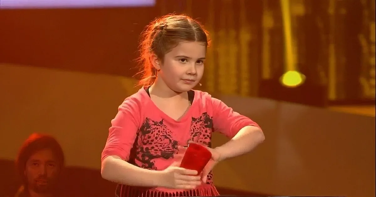 A young girl in a pink dress holds a red object, standing on a stage with a blurred audience and stage lights.