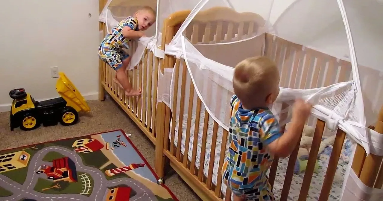 Two toddlers in colorful outfits are climbing out of cribs in a playroom with a toy truck and a colorful play mat.