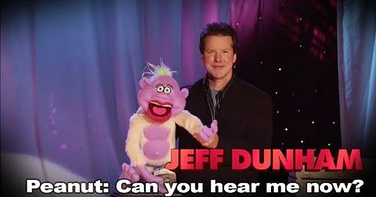 A man in a black suit stands beside a purple puppet with a big smile, against a stage backdrop.