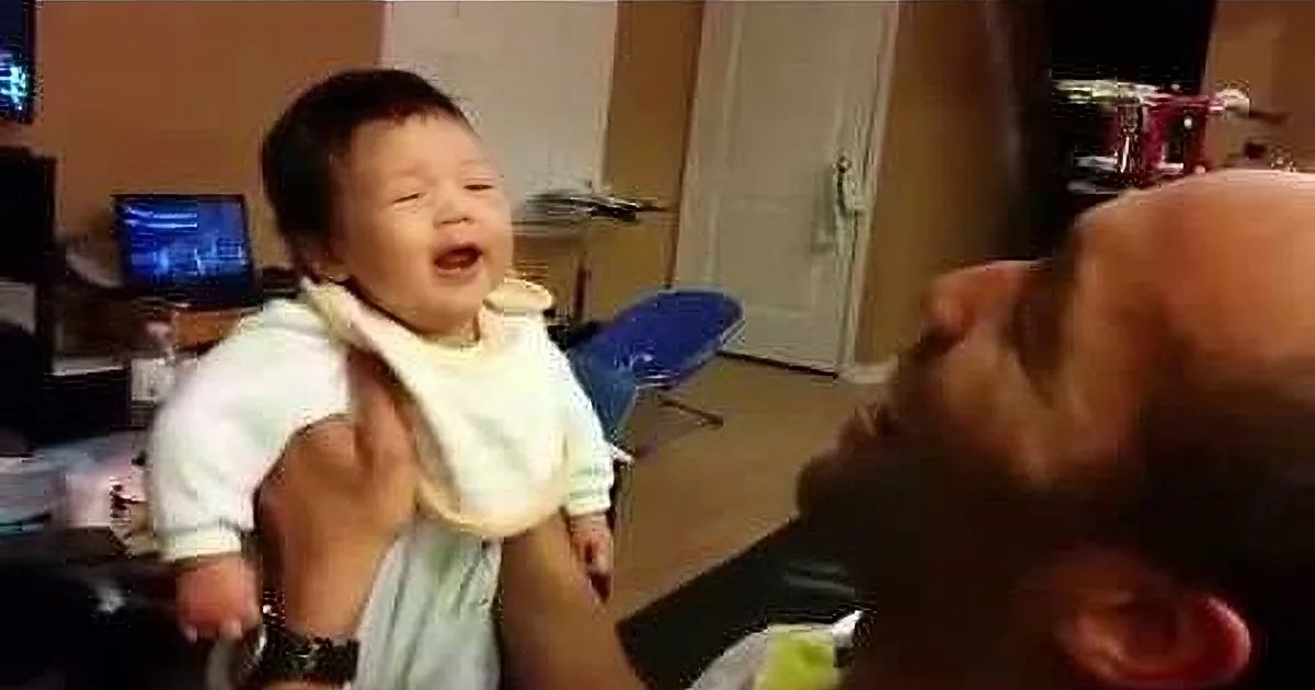 A baby in a bib is being held and appears to be laughing or crying, with a person's hand visible.