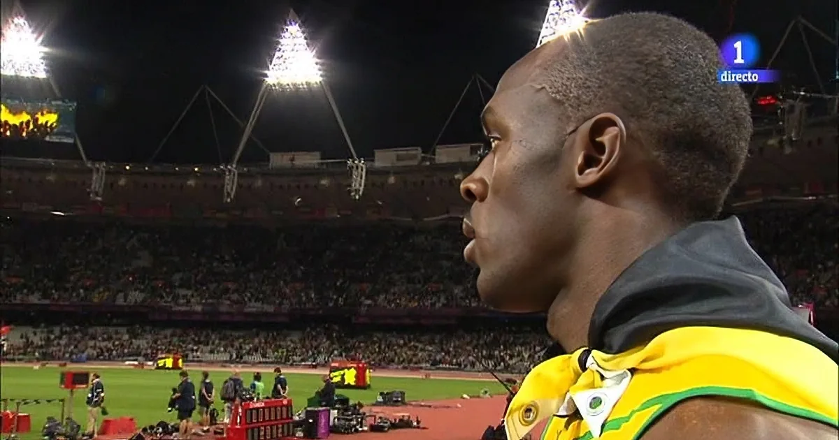 A man in a yellow and green outfit stands on a track, looking towards a stadium filled with spectators.