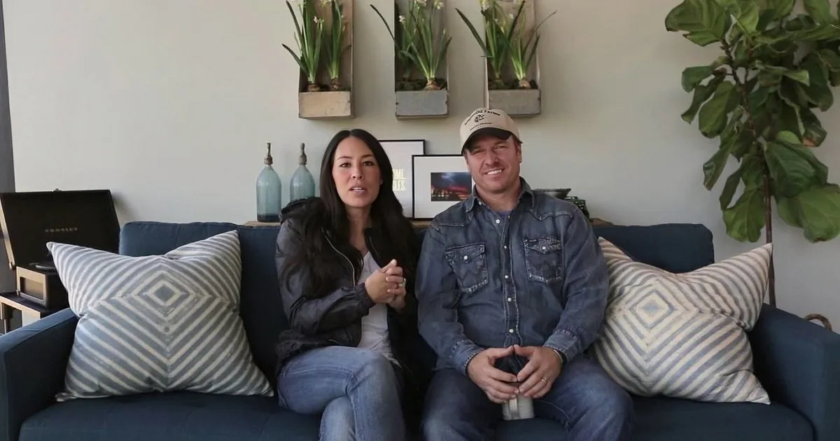 A couple sits on a blue couch with striped pillows, smiling, in a cozy room with plants and decor.