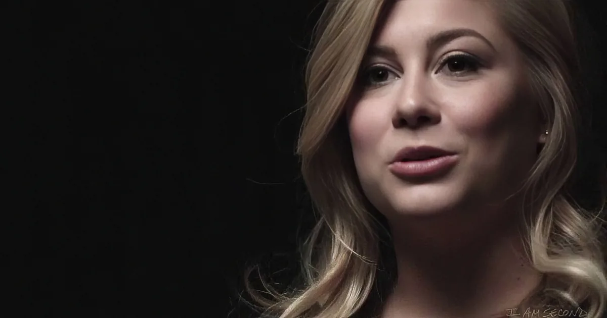 A woman with blonde hair and makeup, wearing a black top, is shown in a close-up against a dark background.