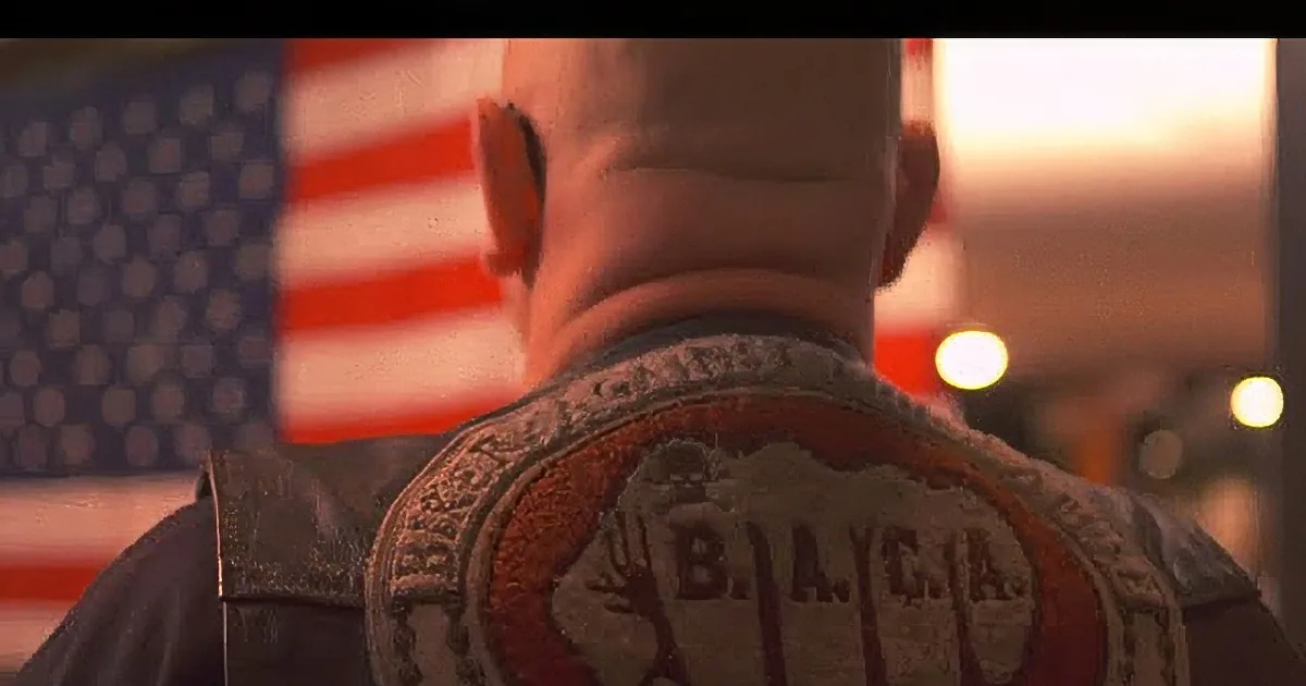 A man with a bald head wearing a leather jacket with patches, standing in front of an American flag.