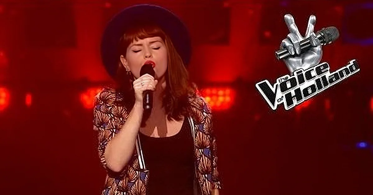 A woman in a hat sings into a microphone on a stage with red lighting.
