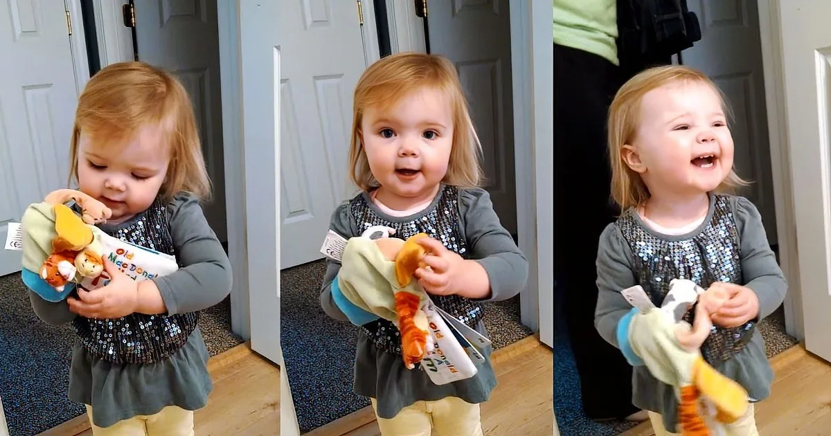 A toddler in a sparkly top and yellow pants holds a stuffed animal, smiling and looking around.