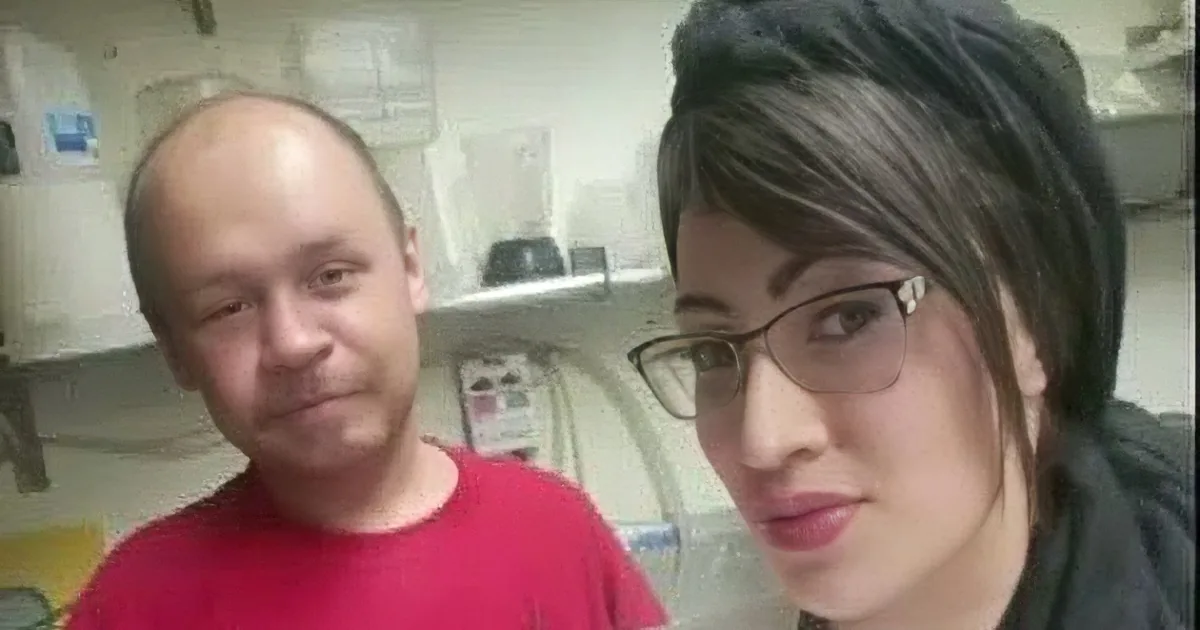 A man in a red shirt is smiling, with a woman in glasses and another person partially visible in the background.