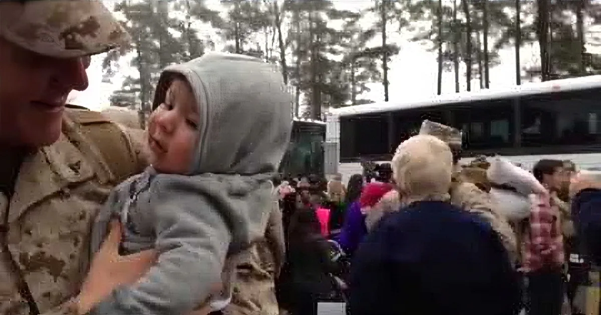 A soldier holds a baby in a crowd near a bus, surrounded by trees.