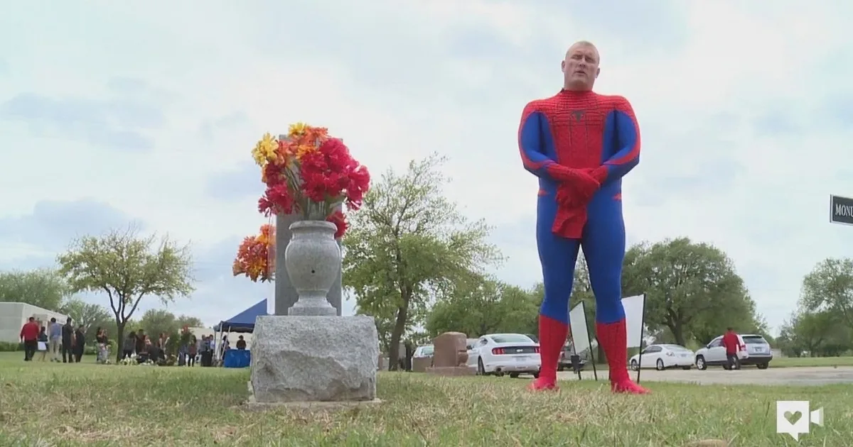 A man in a Spider-Man costume stands beside a grave with flowers, surrounded by a cemetery scene.