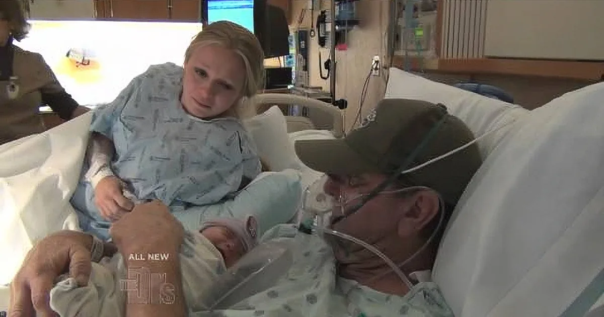 A woman in a hospital bed holds a newborn baby while a man in a cap and oxygen mask looks on.