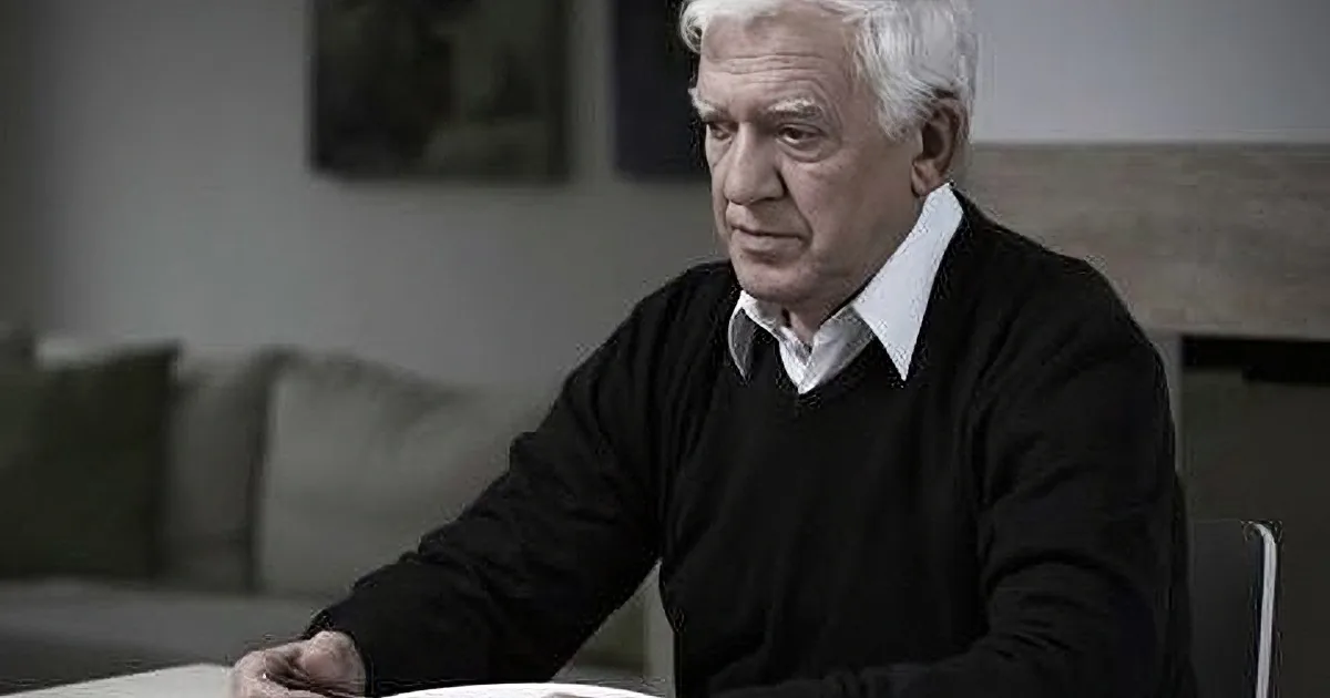 An older man with white hair sits at a table, holding a spoon over a bowl, in a dimly lit room with a couch and artwork...