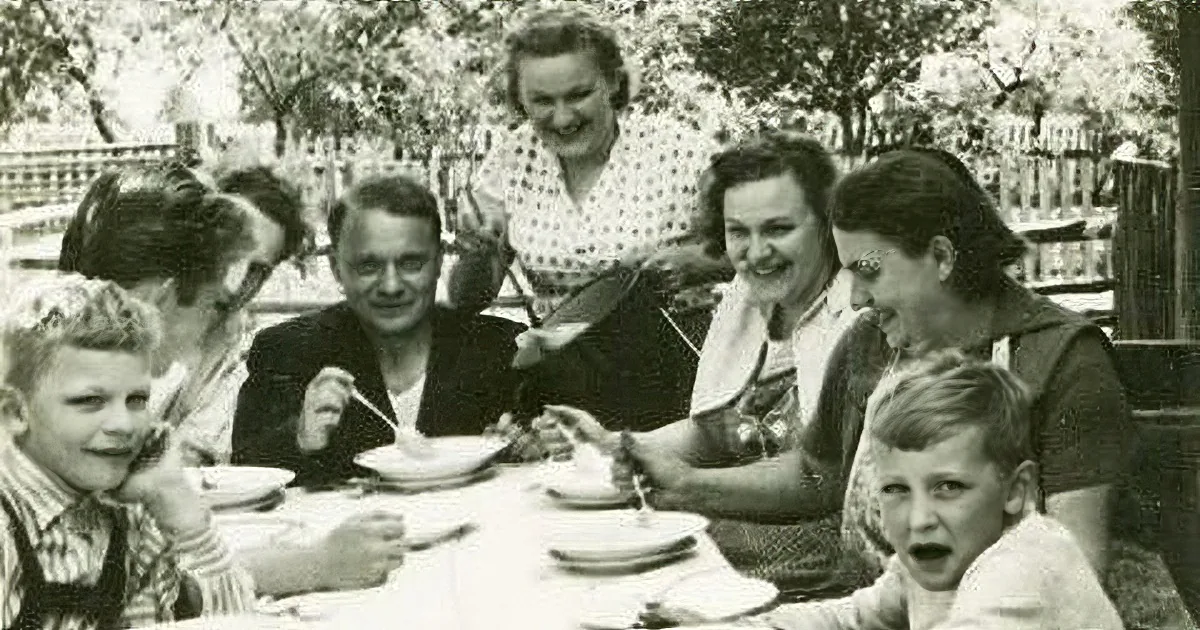 A group of people, including children, are gathered around a table, enjoying a meal together outdoors.