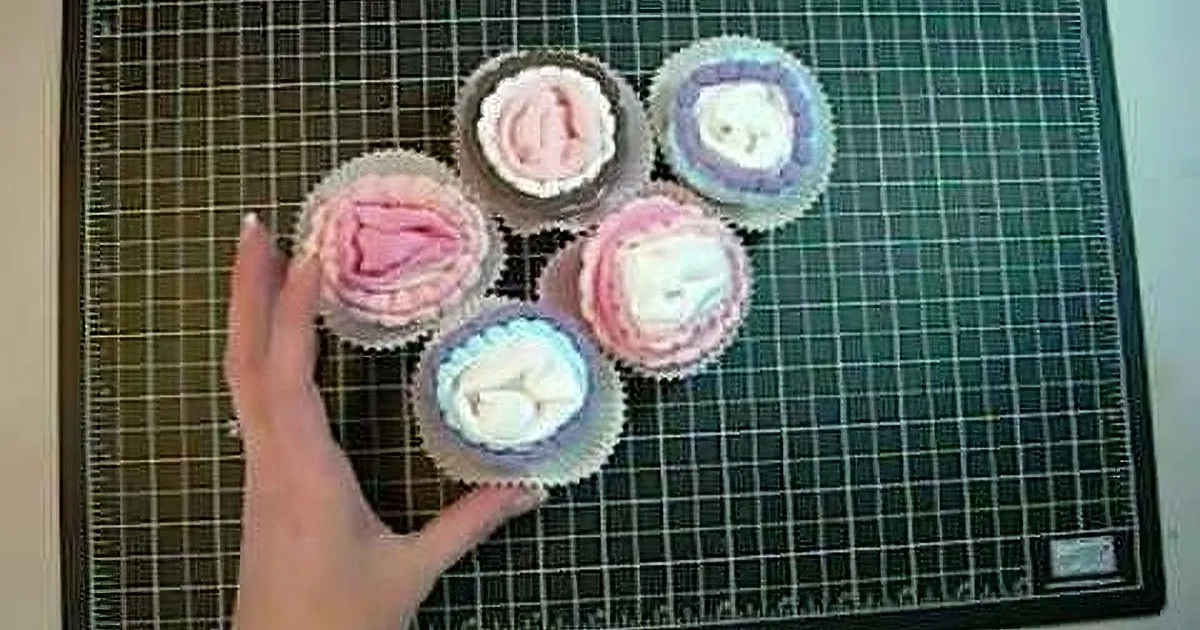 A hand points at a small, decorated cupcake on a grid mat, surrounded by other cupcakes with colorful frosting.