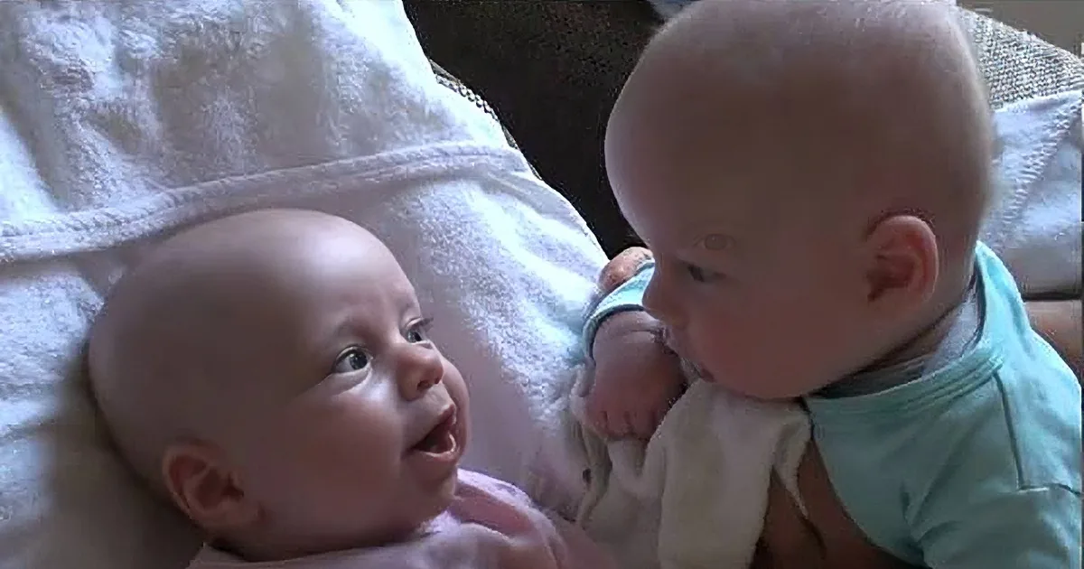 Two babies, one smiling and looking up, the other in a green outfit, both on a white towel.