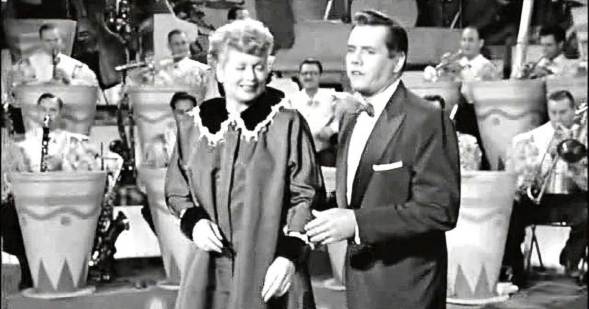 A man in a suit gestures towards a woman in a fur-trimmed coat on a stage with a band.