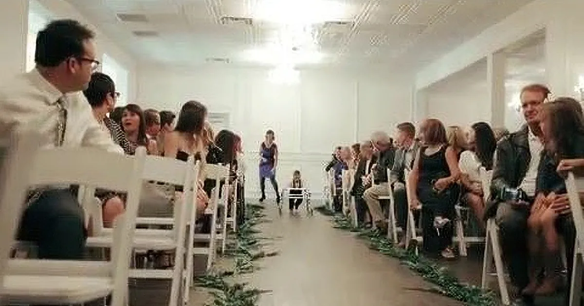 A bride walks down a aisle lined with greenery, surrounded by seated guests in a white-walled room.