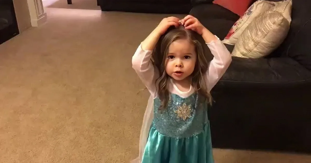 A young girl in a teal dress with a white top is standing indoors, holding her hair up with both hands.