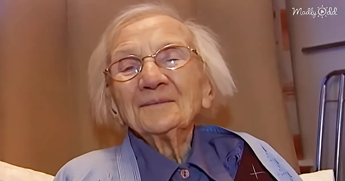 An elderly woman with white hair and glasses, wearing a blue shirt, is seated indoors.