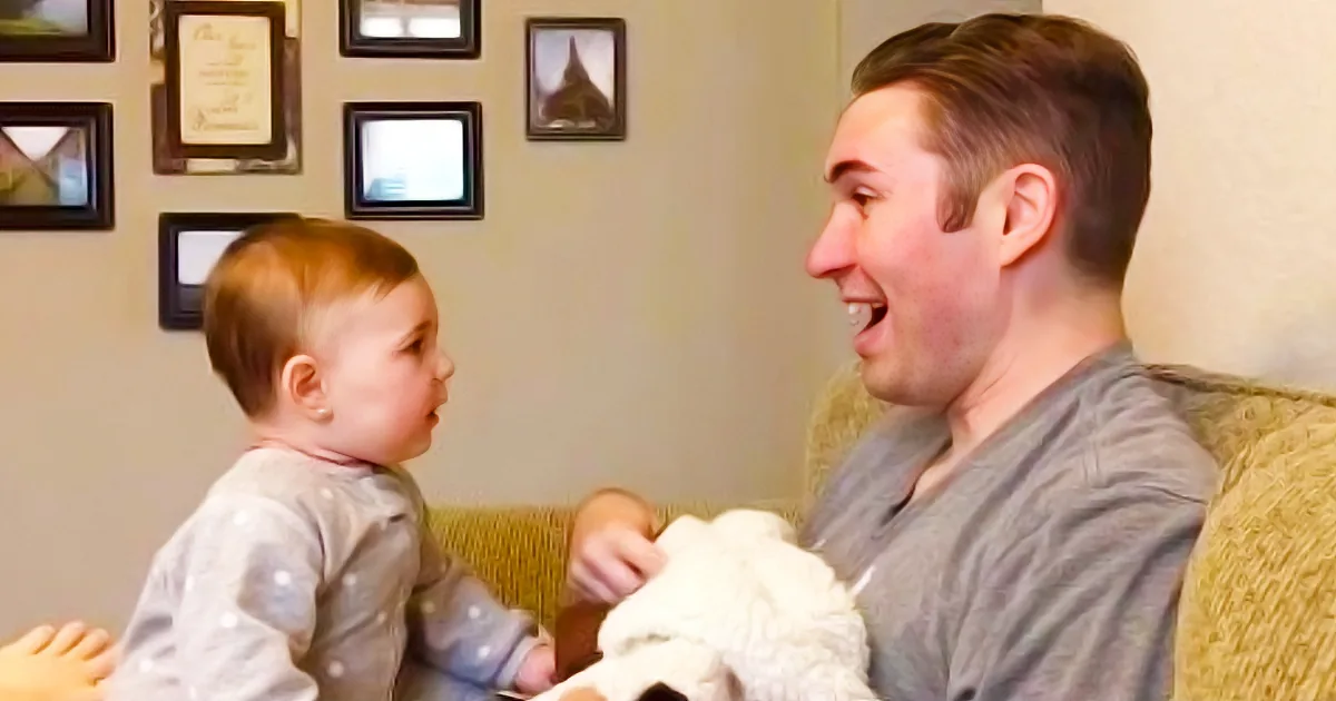 A man and a baby are sitting on a couch, both smiling and looking at each other.