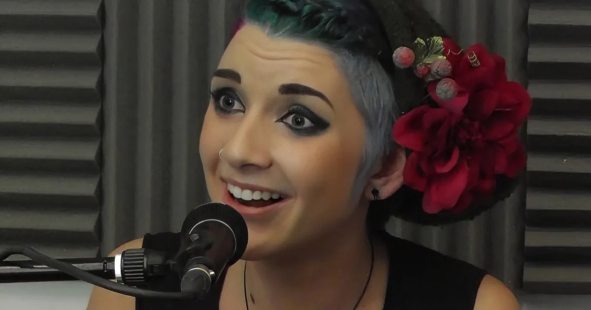 A person with colorful hair, piercings, and jewelry smiles with hands near face, surrounded by festive lights.
