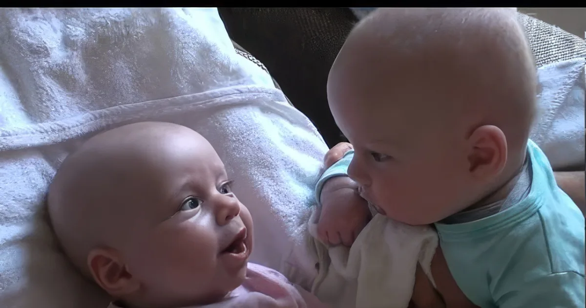 Two babies, one smiling and looking up, the other leaning in close, both on a white blanket.
