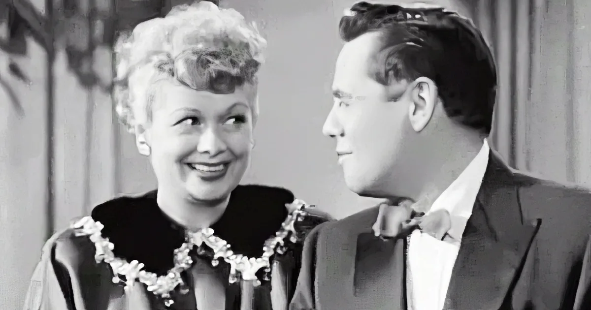 A woman in a black dress with a pearl necklace smiles at a man in a suit.