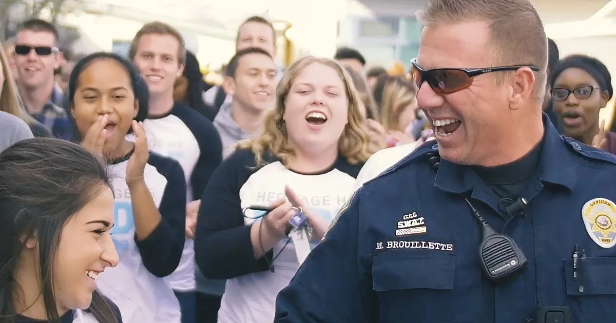 A police officer in uniform smiles and interacts with a group of people, some laughing.