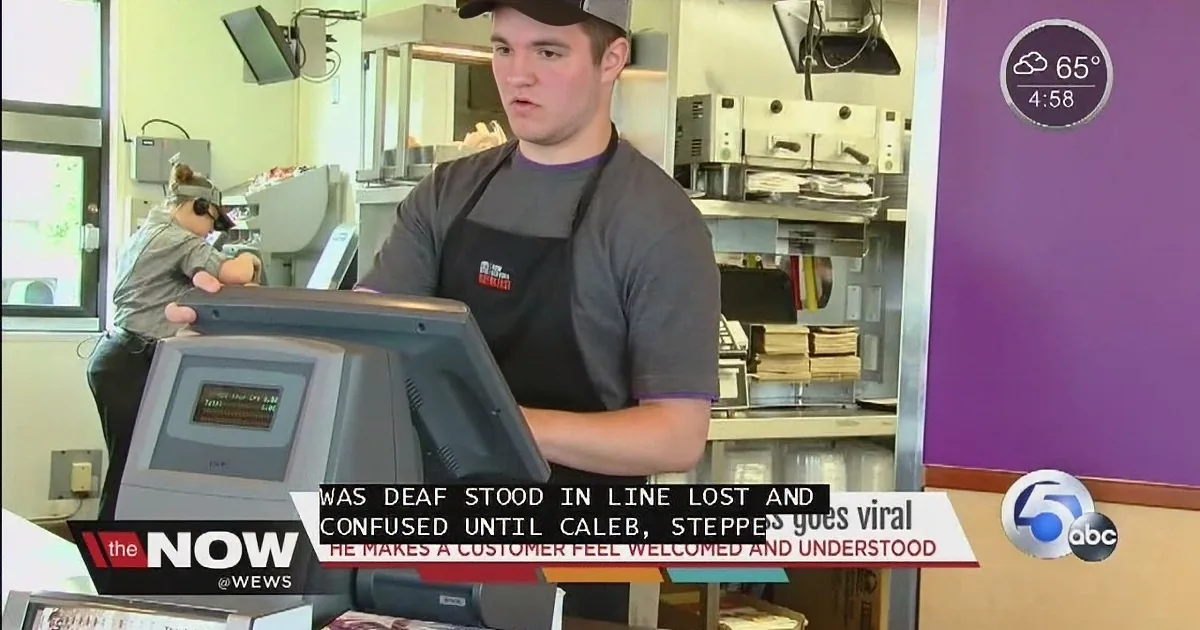 A man in a black apron stands behind a cash register, likely working at a fast-food restaurant.
