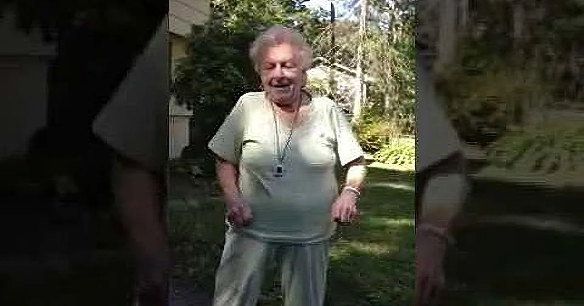 An elderly woman in a light green shirt and beige pants is smiling and walking outdoors, surrounded by trees and greenery.
