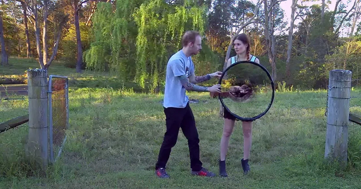 A man and woman are interacting with a large circular object in a grassy outdoor setting.