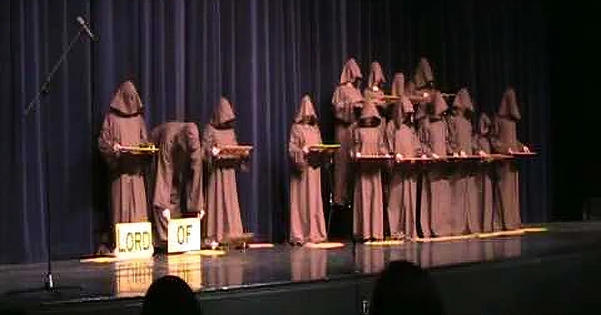 Monks in brown robes stand on stage, holding signs reading "LORD OF" and "OF".