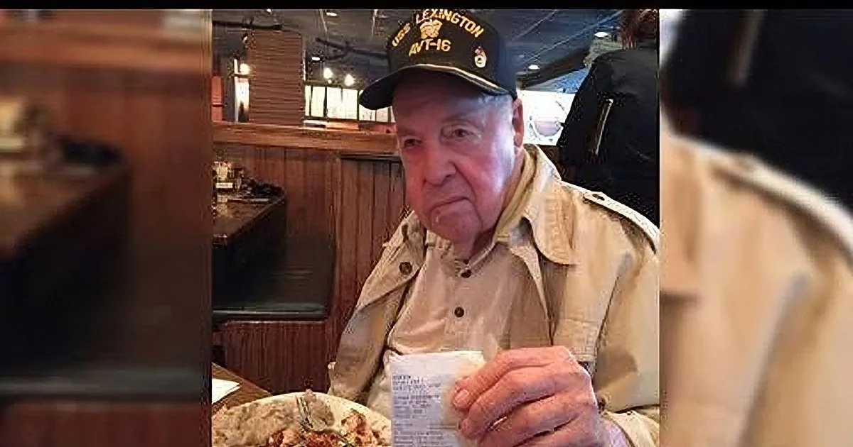 An older man wearing a Navy hat sits at a restaurant table, holding a napkin, with food and a menu in front of him.